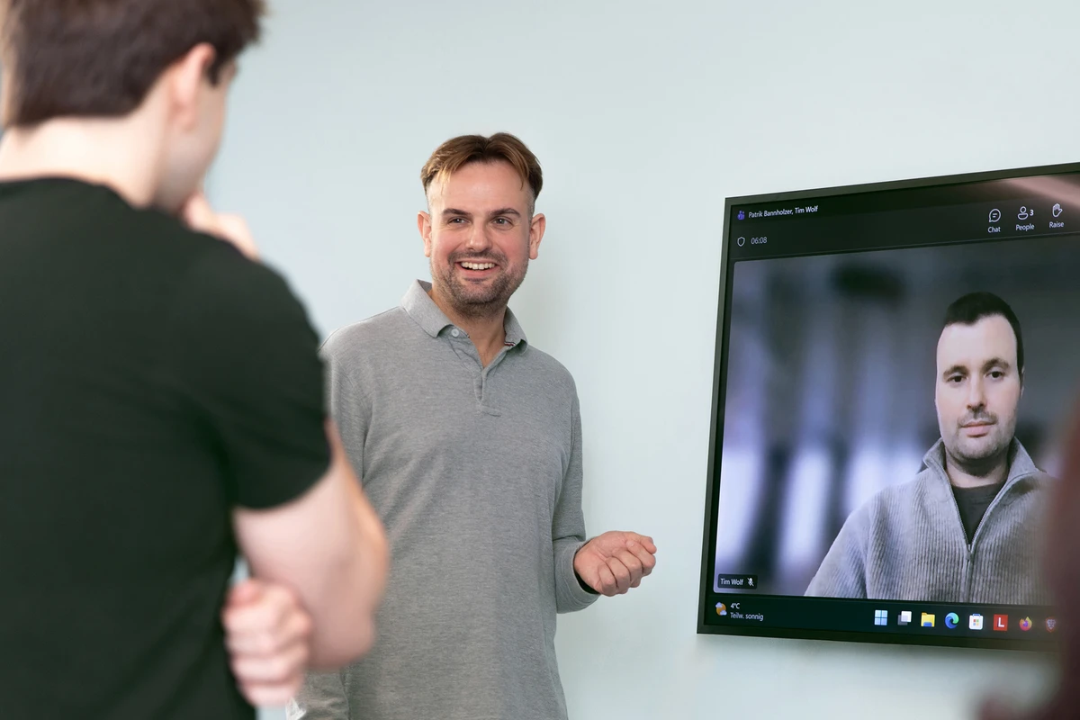 Mann steht vor einem Bildschirm und spricht zu mehreren Personen in einem Raum.