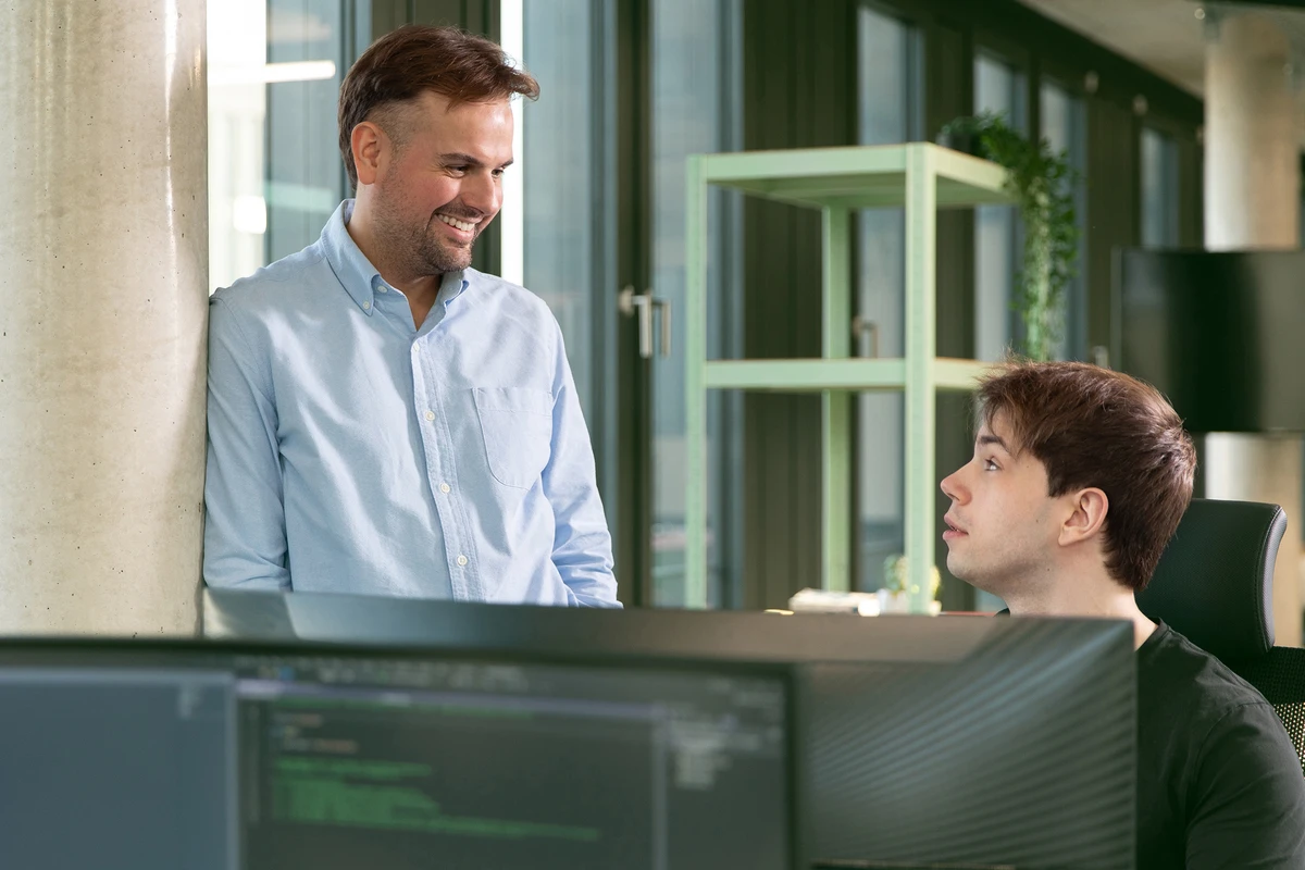 IT-Karriere mit Fokus auf Data Consulting und Teamwork. Eine Person steht und tauscht sich mit seinem Kollegen vor einem Rechner über ein Projekt aus.